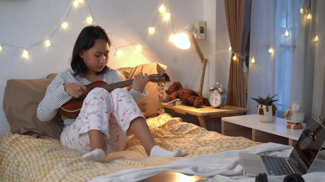 Young girl sits comfortably on her bed practicing the ukulele after her online classes in bedroom illuminated by warm fairy lights