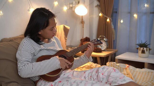 Young girl sits comfortably on her bed practicing the ukulele after her online classes in bedroom illuminated by warm fairy lights