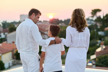 Obraz premium Family on Rooftop Balcony Watching Sunset Over Coastal Town Parents and Son Embracing Togetherness