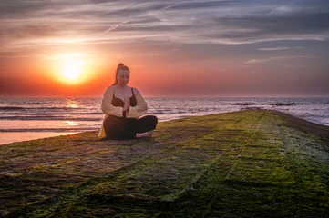 Fototapete Beruhigend Woman meditating with palms pressed together and head gently bowed while seated cross legged on a breakwater at sunset a calming gesture of gratitude and inner peace.  © Oksana