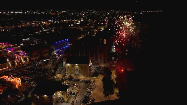 High altitude aerial parallax over Willemstad Curacao captures fireworks bursting above Tempel Emanu-El Synagogue surrounded by glowing cityscape and crowds