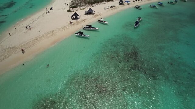 Drone zenith tilt up over crystal-clear turquoise waters reveal one of the best beaches in the world with awnings and tents for visitors and private boats.