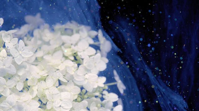 Close-up scene of white hydrangea cluster at left foreground while translucent blue veil flows through midground, with dense multicolored bokeh, glitter, soft light, deep navy background