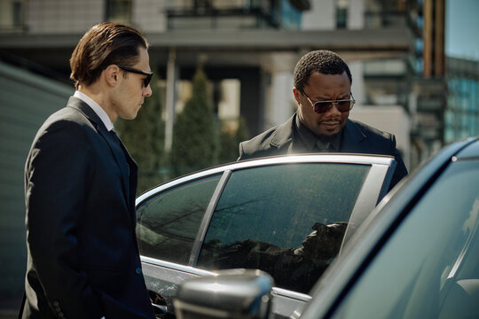 Caucasian man and African American man standing by car, opening door during executive transport service. Ideal for corporate security, VIP travel, business mobility marketing