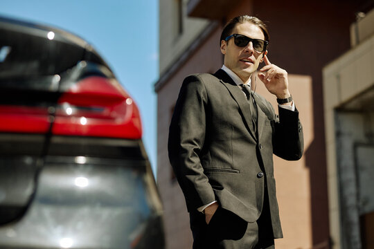 Portrait of young adult Caucasian businessman standing near car, speaking on earpiece and monitoring surroundings. Ideal for executive security, corporate transport, business travel