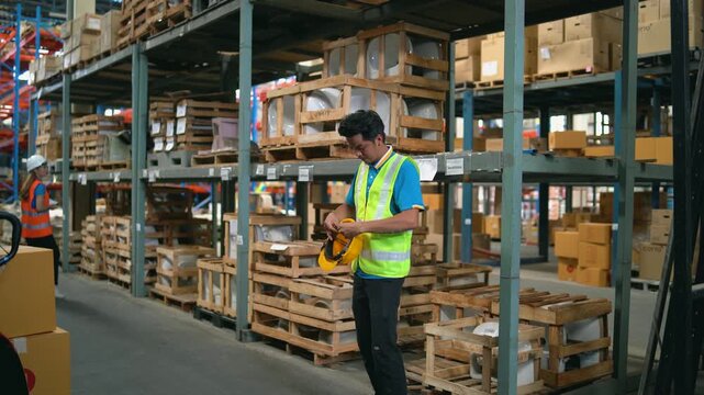 Warehouse worker wearing safety vest inspecting clipboard and holding hard hat surrounded by shelves with stacked cardboard boxes in industrial storage facility