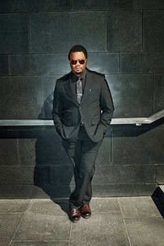 Portrait of African American young adult businessman standing against urban wall, posing in formalwear and looking at camera, conveying leadership and executive branding for corporate marketing
