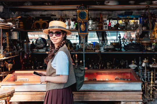 Smiling woman with sunglasses holding a phone while standing inside an antique shop. Horizontal