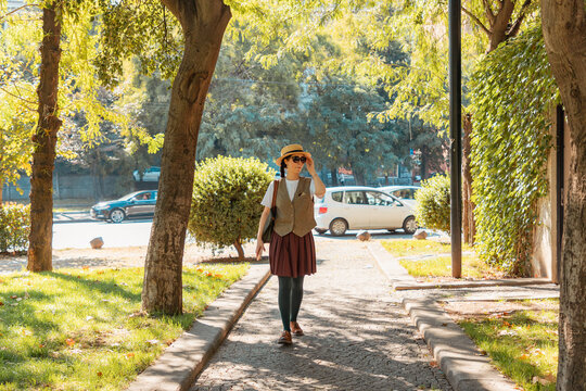 Young woman in a straw hat strolling along a sunlit cobblestone pathway surrounded by trees. Horizontal