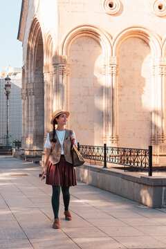 Young woman in a straw hat walking past a stone building with arched columns and a wrought iron fence. Vertical