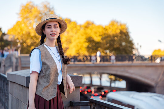 Young woman with braids and a straw hat standing on a bridge with blurred autumn trees and city traffic. Vertical