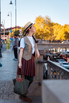 Woman standing on a stone bridge railing holding a bag, looking at the street below. Vertical