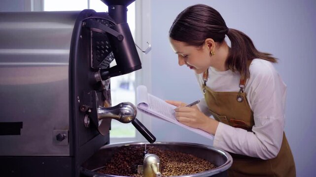Female coffee roaster taking notes on clipboard near roasting machine