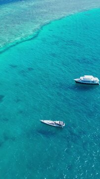 Aerial view yacht anchored turquoise lagoon overlooking coral reef and tiny island panoramic drone shot captures luxury vessel with tender moored nearby, crystal clear shallow,Maldives, vertical video