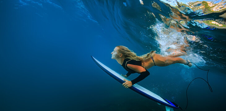 Side View Of Female Surfer Paddling Underwater With Surfboard In Clear Blue Ocean Water