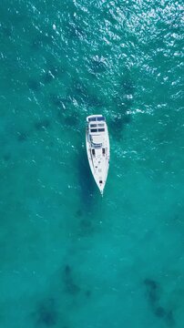 Close aerial sailboat on turquoise lagoon single sailing vessel gliding over clear shallow coral waters captain tending lines sunlight sparkling on hull calm breeze ideal, Maldives, vertical video