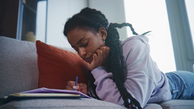 Young person writes in a notebook while lying on a sofa in a cozy indoor space during daylight hours