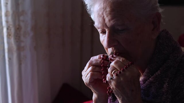 Elderly christian woman holding a rosary and praying with her eyes closed, seeking comfort and spiritual guidance with a small candle lit in front of her, symbolizing her deep devotion and faith