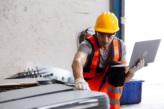 Construction worker in safety gear using a laptop to inspect stacked metal sheets in a warehouse, combining industrial labor with digital quality control.