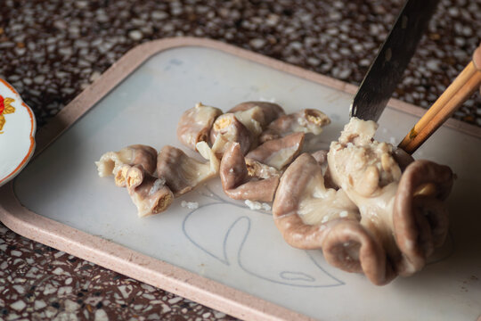 Close up of hands using a knife and chopsticks to slice boiled pork intestines on a white cutting board