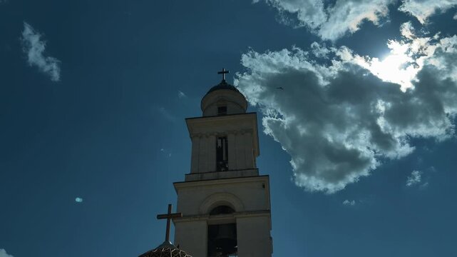 Low-angle view of church bell tower and rooftop cross over historic stone facade with bright cloud passing across sunlit sky, creating backlight and rim light, with church, tower, cross, dome detail