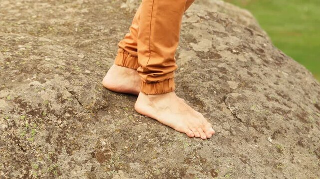 Bare feet balance carefully on a mossy rock surface. Brown pants flow down the legs. Green grass forms a blurred backdrop. Calm focus suggests natural movement