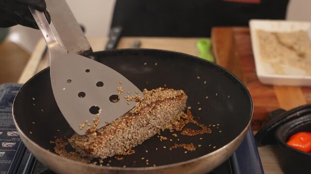 Cook uses a spatula to flip sesame-crusted salmon teriyaki in a frying pan on a stovetop, showing texture and cooking process for recipe or cooking tutorial content.