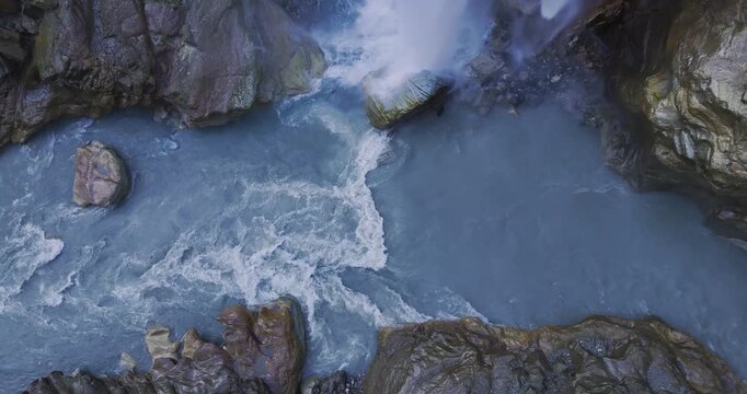 Annapurna Circuit, Nepal, aerial pull-out shot of Danaque waterfall plunging from a rocky cliffside into the winding Marsyangdi River gorge, surrounded by forest trees in the Manang region.