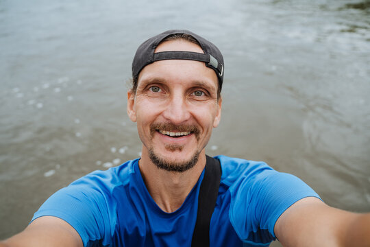 Smiling man in backward cap taking selfie beside river shoreline under soft daylight, relaxed outdoor portrait with rippling water, casual travel mood, natural summer adventure