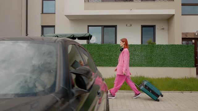 A woman in a pink outfit walks along a path with a suitcase near a block of flats. She is heading out for travel or vacation with her luggage.