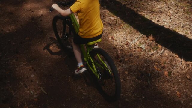 Slow motion elevated rear left view of a school age boy riding a bicycle along a sunny pine forest trail, moving farther ahead and leaving the frame at the end of the summer ride. High quality 4k