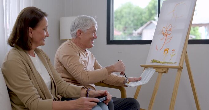 Senior man paints flowers on canvas while senior woman checks tablet reference beside him at home, blending creative hobby, companionship, mindful relaxation, and active retirement wellness.