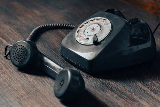 Vintage black rotary dial telephone with a detached handset on dark wooden table, closeup. Antique communication device with dust showing its age, perfect for retro concepts