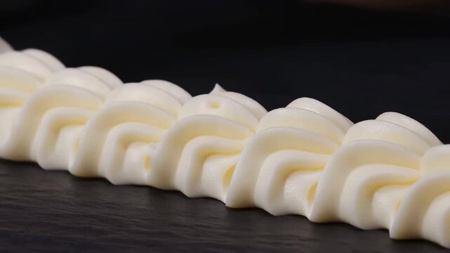Close-up piping buttercream rosettes on dark slate with stainless steel nozzle creating repeating ridged swirls and pointed peaks in decorative pastry detail, with buttercream, frosting, piping