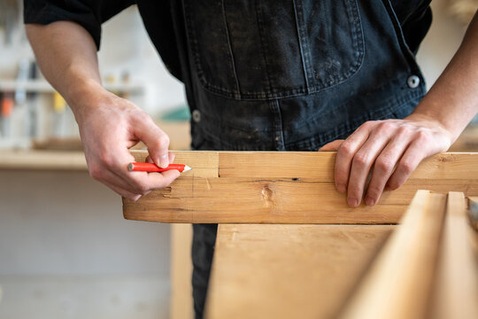 Woodwork specialist processing with wooden boards at worktable, preparing details and marking planks, master in workshop checking fitting of wooden patch for window frame, drawing with pencil on board