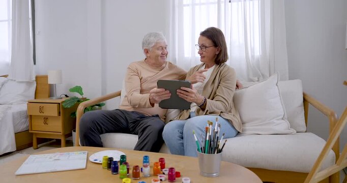 Retired couple sit beside art supplies, scrolling tablet and discussing visual references before painting, sharing laughter, companionship, creative planning, cognitive engagement, and home wellness.