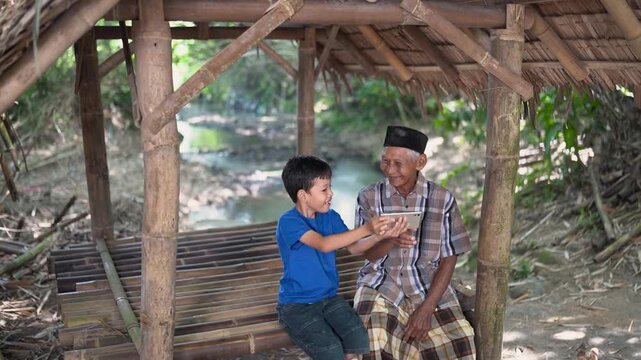 Young boy stands beside smiling elderly man beneath rustic bamboo shelter, both focused on smartphone in shared hands, surrounded by woven roof, wooden platform, calm water, and soft daylight