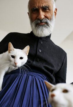 Stylish charismatic Indian, Egyptian or Middle Eastern man in avant‑garde fashion looking down at camera in dramatic low‑angle portrait with two white cats, pleated navy trousers & buttoned black top
