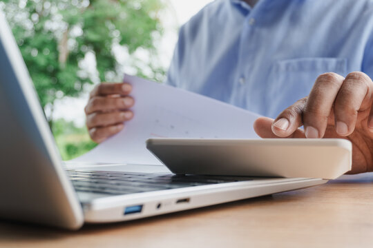 Business professional calculating financial data with calculator while reviewing documents and working on laptop in a natural workspace, representing budgeting, accounting, financial planning