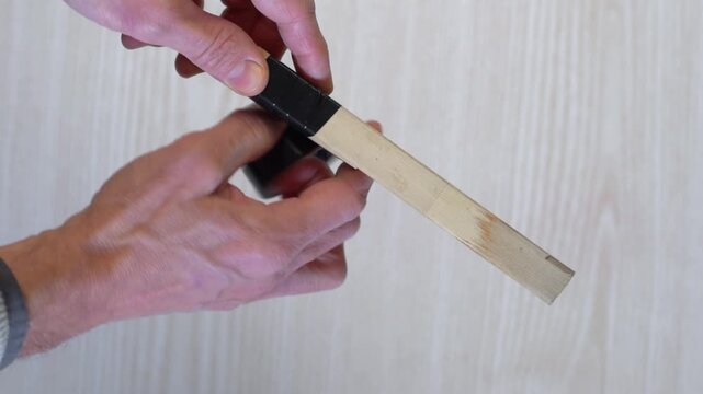 Hands demonstrating taped wood repair closeup, stepbystep demo of wrapping black electrical tape around thin wooden strip on pale table, visible wrist movement and practical