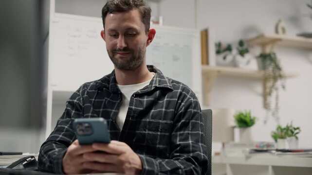 Happy Caucasian man programmer smiling and using smartphone at office desk.