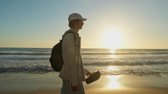 Happy young man walking on a sandy ocean beach on a sunny summer day, in slow motion. Young Male guy walking along the sandy coast on a sunny evening. A guy with a backpack walks on a beach