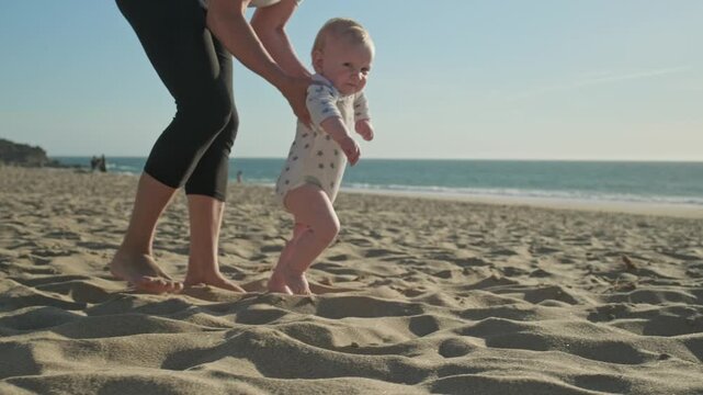 Mom teaches her baby to walk on a sandy sea beach. Toddler takes the first steps with caring mother. Kid takes first steps with the support of his mother on nature. Baby is learning to walk, outdoors.