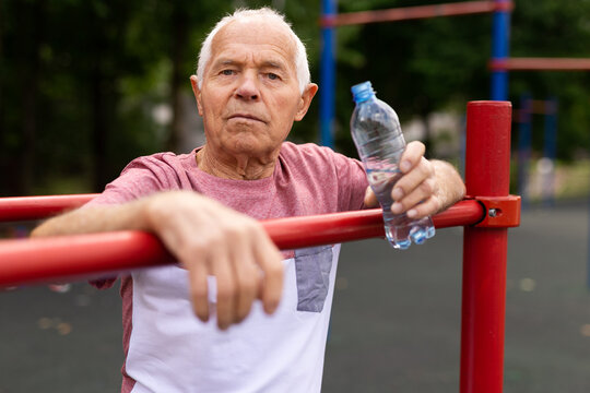 Old man with bottle of water standing in outdoor workout station