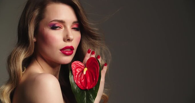 Fashion model with  hair blowing in the wind. Sexy blonde woman with red lips holds a red flower. Closeup face of a pretty young woman with bright makeup.  Slow motion.