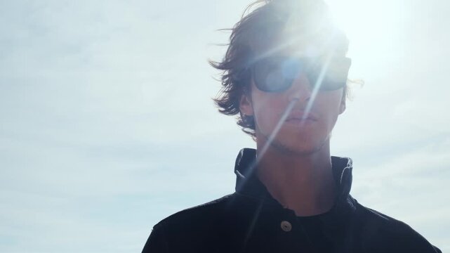 Handsome guy with long hair poses against sky with  sun rays. Stylish man in black jacket looks at the camera, low angle shot. Young man with blown brown hair against the blue sky on summer sunny day.