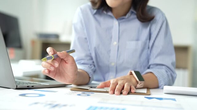 Unrecognizable business woman analyzing data on documents with charts and graphs, working on a laptop computer and taking notes during an online video conference call