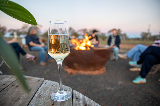 Glass of sparkling wine on a wooden table, with a crowd of women around a firepit in the background