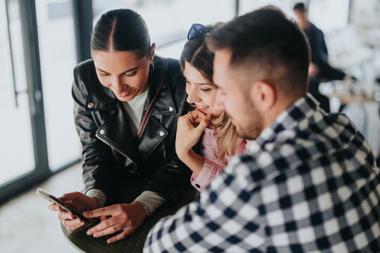 Three friends gathered around a smartphone, smiling as they look at the screen. They share a moment of conversation and amusement while seated in a bright cafe.