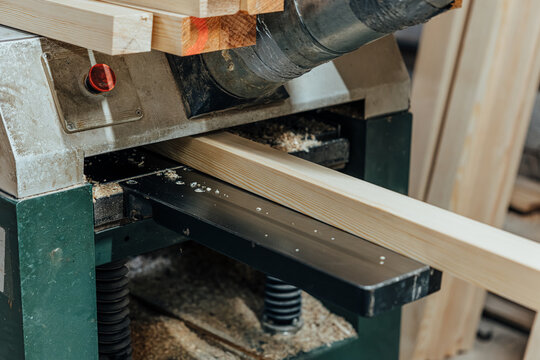 Close-up of a softwood beam moving through a woodworking thickness planer, with visible grain, wood shavings, dust extraction pipe, and workshop machinery